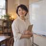 松本奈緒老師的示意肖像：短髮、淺色襯衫，拿著筆記本站在空白白板旁（AI 生成的示意照片）
